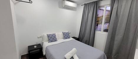 A hotel room with a bed, a nightstand, an air conditioning unit, a window with curtains, and a view of the outside.