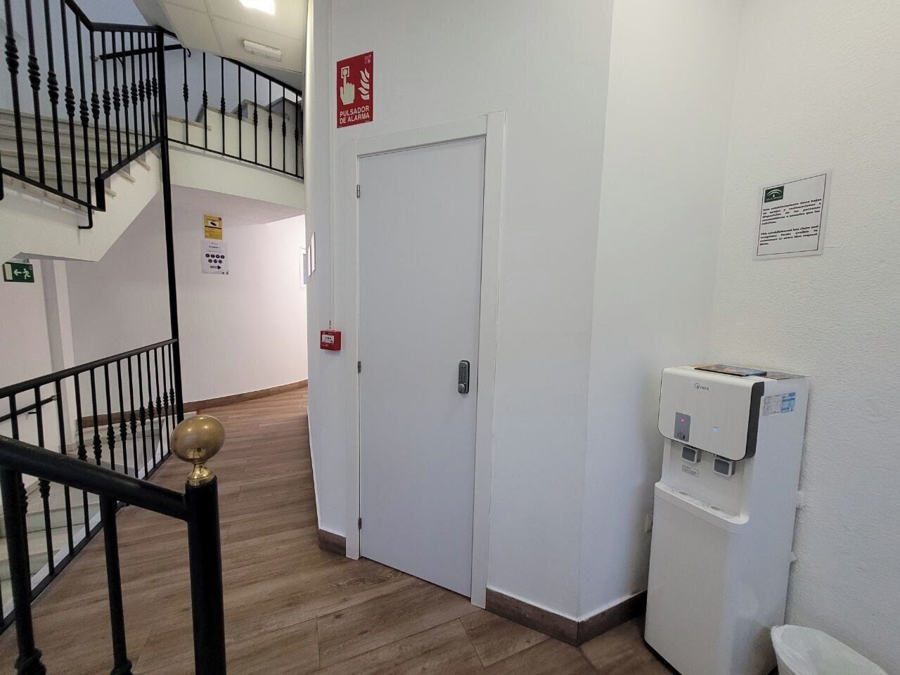 Un corridoio con una porta bianca, un dispenser d'acqua e una scala con ringhiere nere.