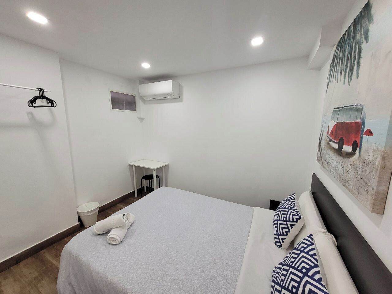 Une chambre bien rangée, avec un lit, des oreillers, une petite table, une chaise, un appareil de climatisation et un porte-manteau fixé au mur.