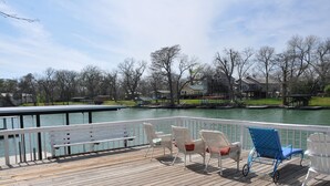 A wooden deck with white and blue chairs, a bench, and a view of a lake with houses in the background. - Large Mediterranean pool, river, basketball, sleeps 26, 8 bedrooms (seguin)