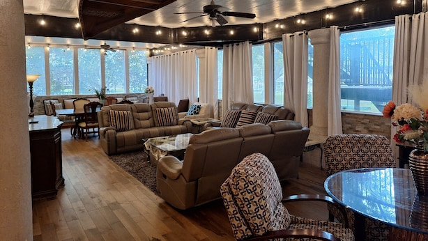 A spacious living room with a large sectional sofa, a glass coffee table, and a dining area with a table and chairs. There are multiple windows with curtains, providing natural light and views of the outdoors. - Large Mediterranean pool, river, basketball, sleeps 26, 8 bedrooms (seguin)