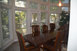 A dining area with a large wooden table and six chairs, large windows with a view of trees, and a potted plant on the right.