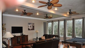 A living room with a pool table, leather sofa, ceiling fans, and a large window with curtains.