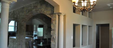A spacious living area with a stone archway leading to a kitchen, a dining table with chairs, and a chandelier.