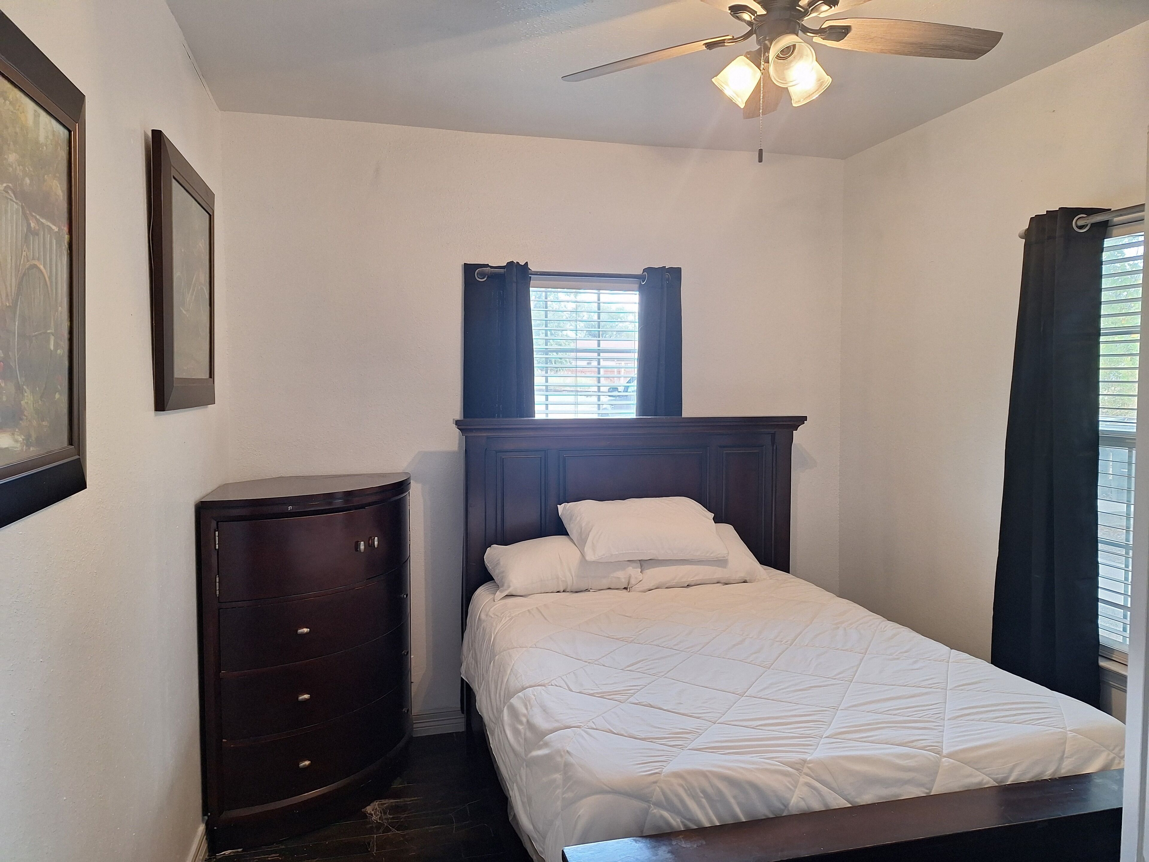 Ein Schlafzimmer mit Bett, Kommode, Fenster mit Jalousien und Deckenventilator.