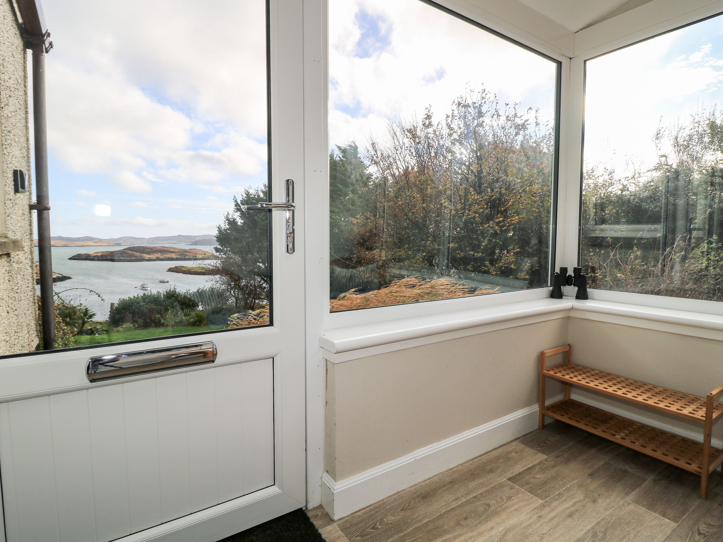 Habitación con un ventanal amplio que ofrece vistas a un cuerpo de agua y colinas. Hay un banco y una estantería con objetos.