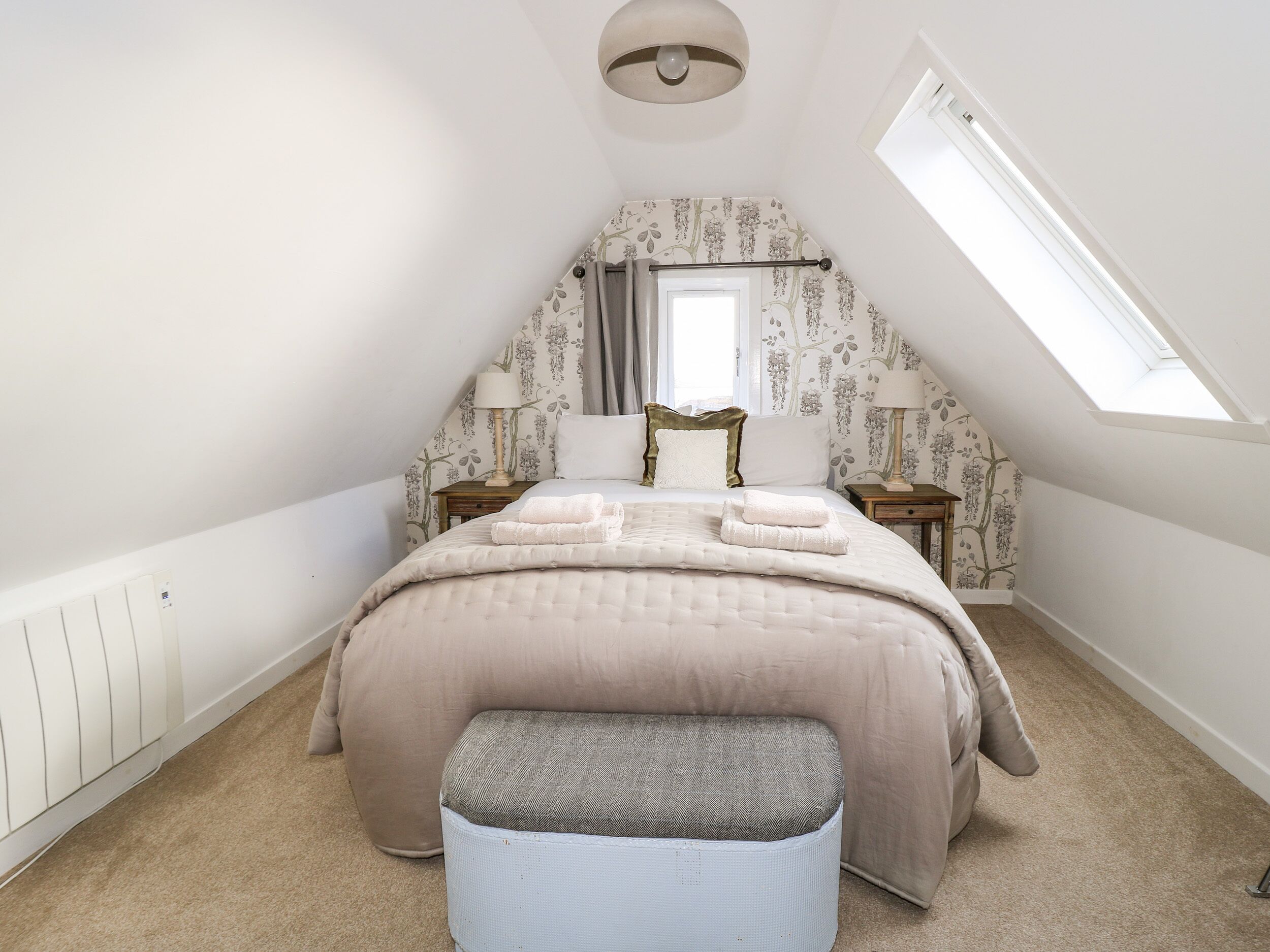 A bedroom with a sloped ceiling, a bed with a beige duvet and pillows, a bedside table with a lamp, a window with a view, and a radiator on the wall.