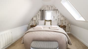 A bedroom with a sloped ceiling, a bed with a beige duvet and pillows, a bedside table with a lamp, a window with a view, and a radiator on the wall.
