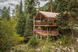 A two-story wooden cabin with a balcony, surrounded by dense forest and mountains. - Creekside Hidden Gem | Close to Downtown Ouray | Two Decks | STR2025-028 (Ouray)