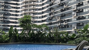 A swimming pool with a tiled floor, surrounded by lush greenery and a modern apartment building with balconies in the background. - One Bedroom Balcony With Great Location and Value (Makati)