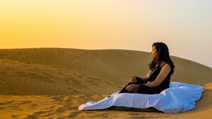 A person sitting on a sand dune with a white cloth, surrounded by sand dunes and a sunset in the background. - Marvin Desert Camp (Jaisalmer)