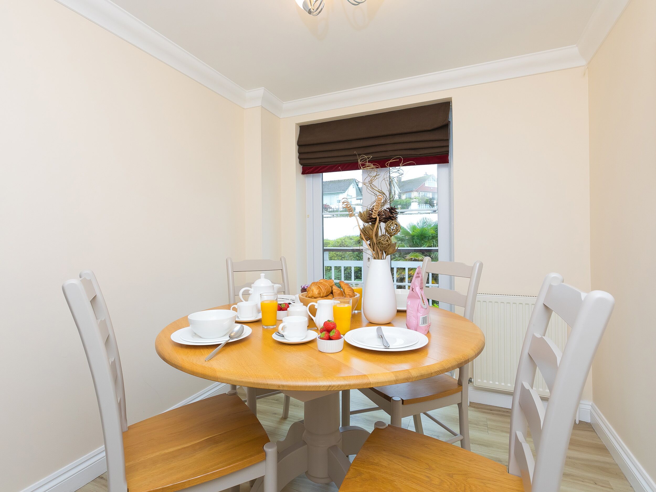 A dining area with a round wooden table, white chairs, and a window with a view of greenery and buildings.