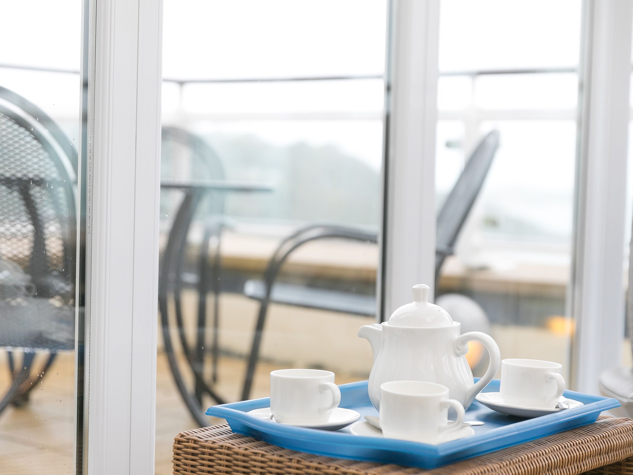 A breakfast setup with a teapot, cups, and saucers on a tray, placed on a wicker table near a window overlooking a balcony.