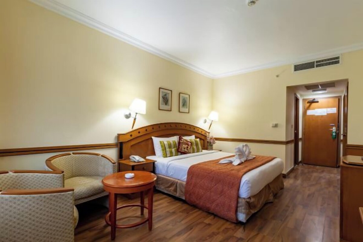 A hotel room with a bed, two chairs, a small table, and a bedside lamp.