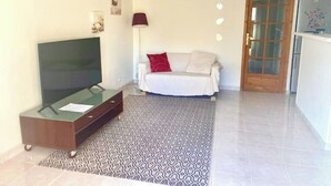 A living room with a flat-screen TV on a wooden cabinet, a sofa with a red pillow, a patterned rug, and a wooden door. - Deux Pièces Avec Terrasse et Parking au Calme (Cagnes-sur-Mer)