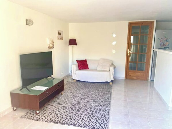 Una sala de estar con una televisión de pantalla plana sobre un mueble de madera, un sofá con un cojín rojo, una alfombra estampada y una puerta de madera.