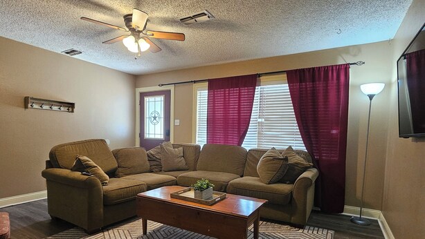 Ein Wohnzimmer mit einem Ecksofa, einem Couchtisch, einem gemusterten Teppich, einem Deckenventilator und einem Fenster mit Vorhängen.