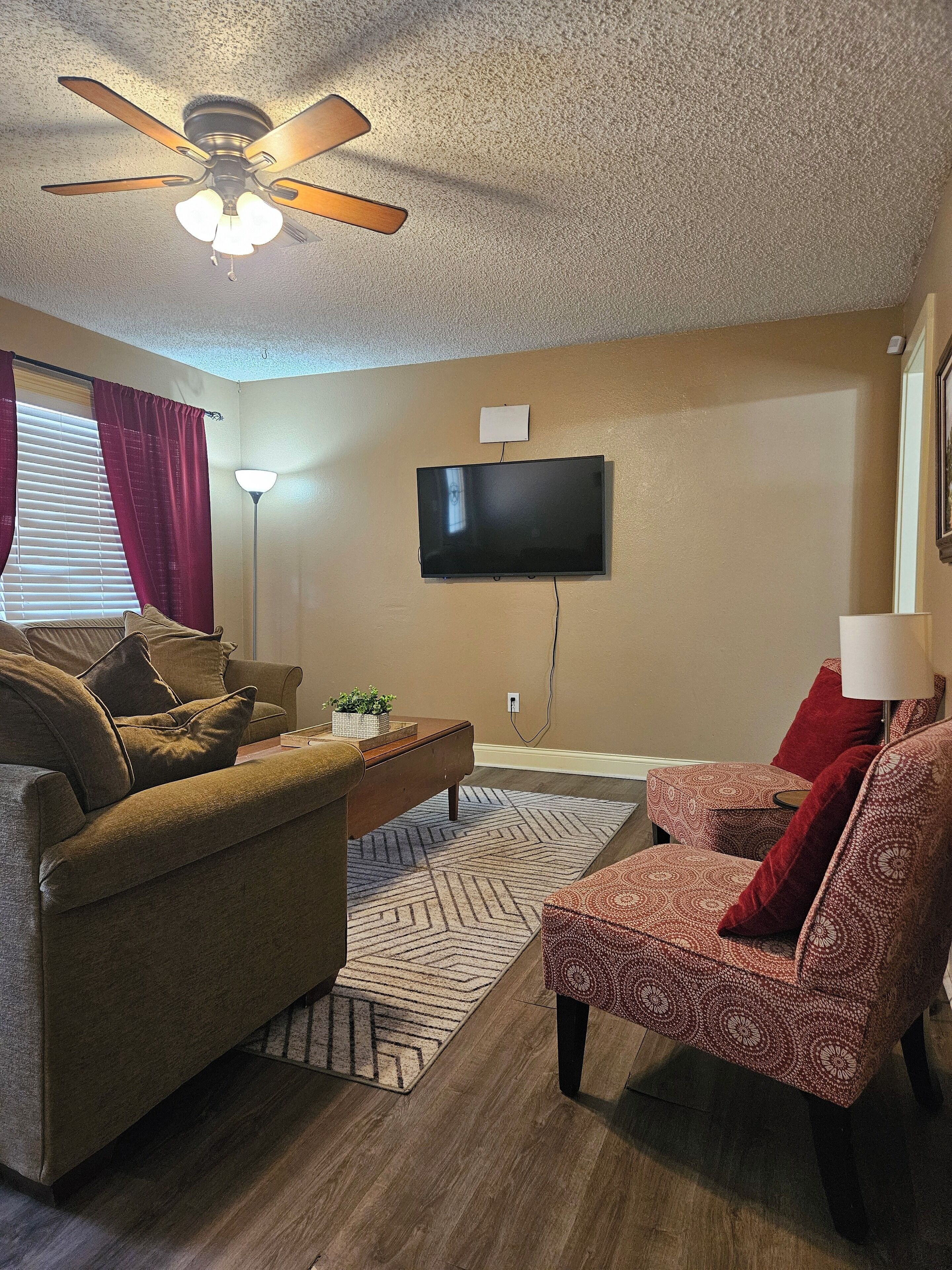 Un salon doté d’un ventilateur de plafond, d’un téléviseur à écran plat fixé au mur, d’un canapé, d’un fauteuil à motifs et d’une table basse.
