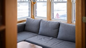 Ein Zimmer mit grauem Sofa, einer Lampe und einem Fenster mit Blick auf eine verschneite Landschaft.