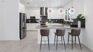A modern kitchen with white cabinets, a stainless steel refrigerator, a black tiled backsplash, and three bar stools. - Sunshine Shores | Beach-facing Stunner w/ Sunny PVT Pool, Fairytale BR, Karaoke (Melbourne Beach)