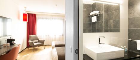 Una habitación de hotel moderna con una cama grande, un escritorio con una silla, un baño con lavamanos y espejo grandes, y una pared de acento roja.