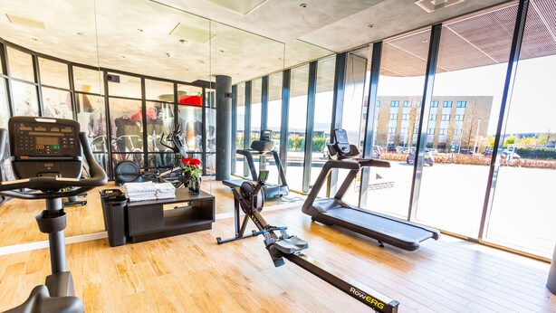 A modern gym with cardio equipment, a reception desk, and large windows offering a view of the outside. - Bastion Hotel Amersfoort (Amersfoort)