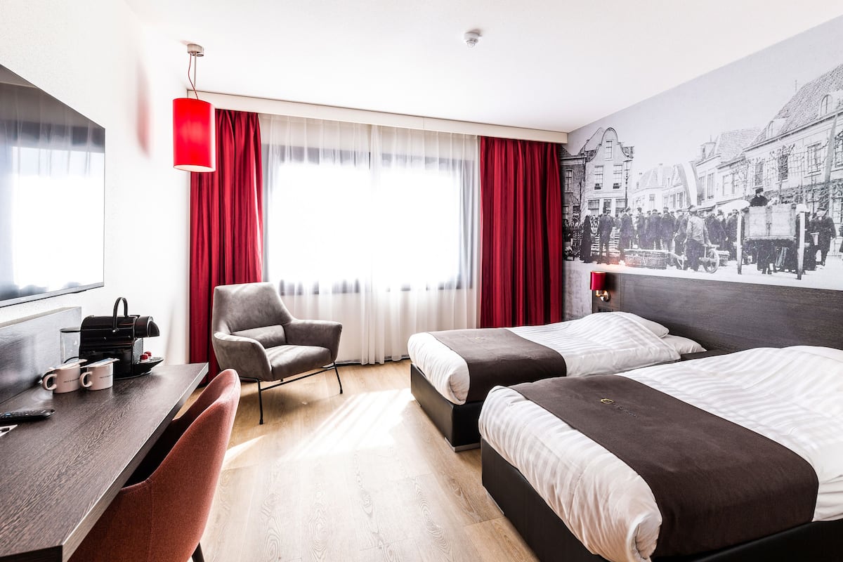Quarto de hotel com duas camas, uma escrivaninha, uma cadeira e uma janela grande com cortinas vermelhas.