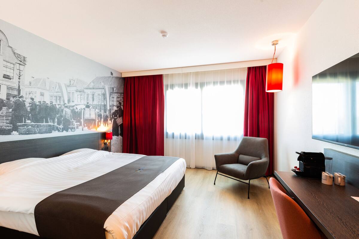Quarto de hotel com uma cama grande, uma escrivaninha, uma cadeira e uma televisão.