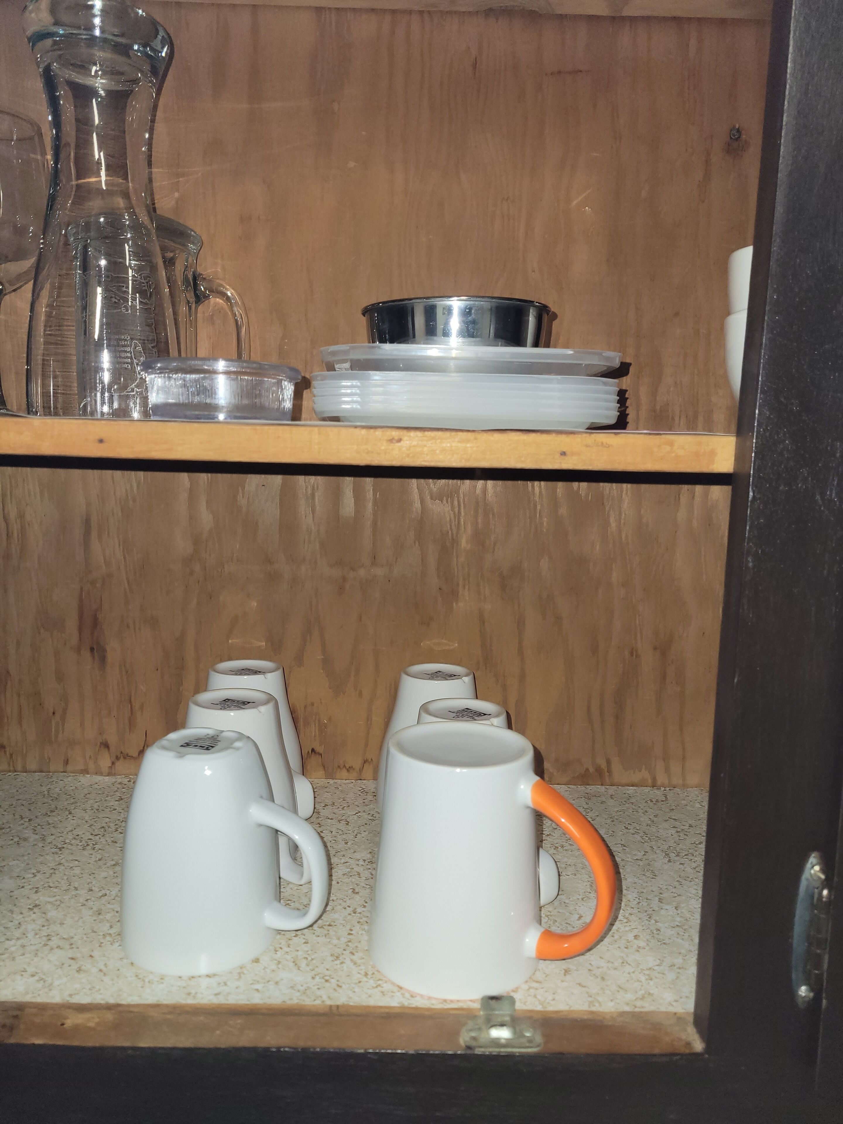 A kitchen shelf with various glassware, including a pitcher, a metal bowl, and several white mugs with orange handles.