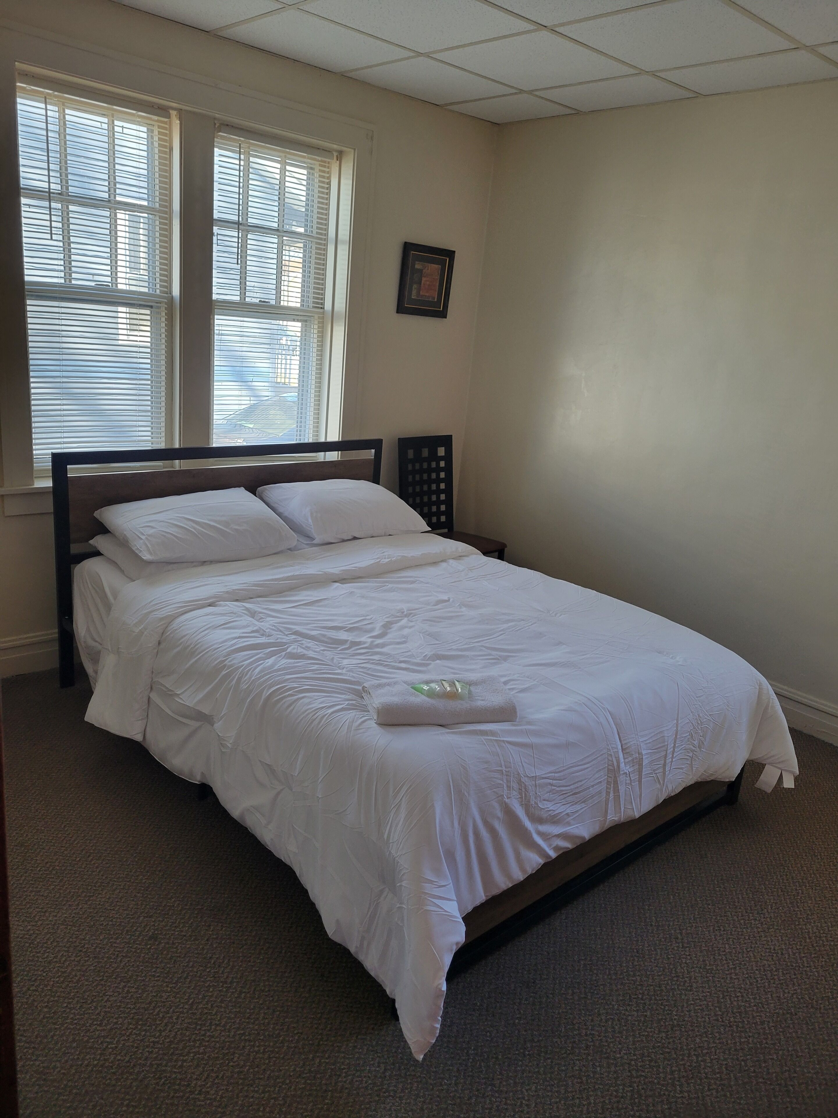 A neatly made bed in a room with a large window, a framed picture on the wall, and a carpeted floor.