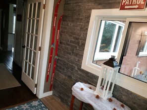 A hallway with a white door, a red sign, a brick wall, a window with a view, and a small table with pine cones. - Beautiful waterfront cottage (with hottub), in the heart of Muskoka. (Huntsville)