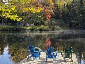 A wooden dock with three Adirondack chairs facing a calm lake, surrounded by trees with autumn foliage. - Beautiful waterfront cottage (with hottub), in the heart of Muskoka. (Huntsville)