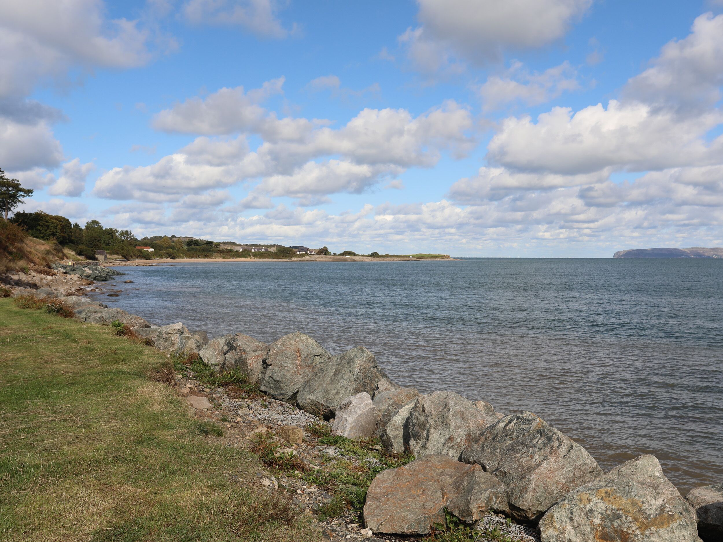 A coastal landscape with a rocky shoreline, calm water, and a distant view of houses on the horizon.