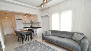 A modern kitchen with white cabinets, a dining table with chairs, and a grey sofa with patterned cushions. - DUYGU APART (Fethiye)