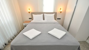 A neatly made bed with a grey bedspread and two white towels, two wall-mounted light fixtures, and a window with sheer curtains. - DUYGU APART (Fethiye)