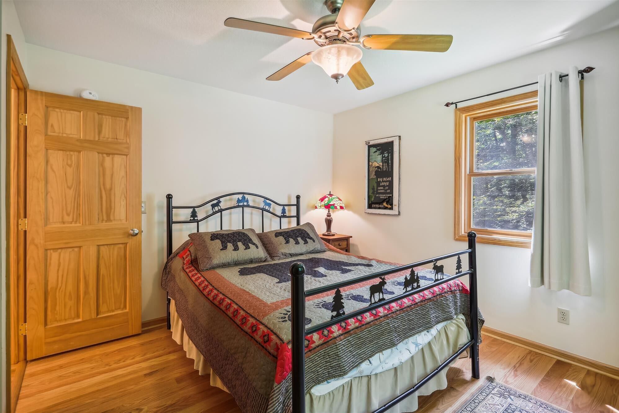A bedroom with a bed, a ceiling fan, a window with curtains, a door, and a poster on the wall.