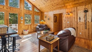 Un interior de cabaña acogedor con paredes de madera, ventanas amplias, un sofá de cuero, un comedor y una chimenea.