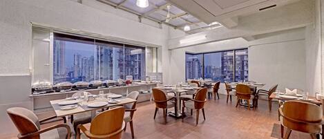 A modern dining area with multiple tables and chairs, large windows with city views, and a high ceiling with exposed beams and lighting fixtures.