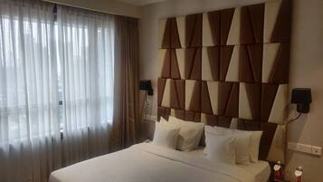A hotel room with a large bed, a headboard with a geometric pattern, two bedside lamps, and a window with sheer curtains.