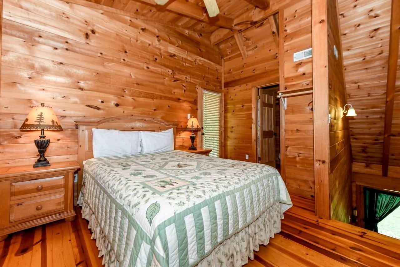 Un dormitorio de cabaña de madera con cama, mesitas de noche y una ventana con persianas.