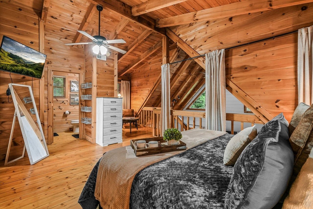 A cozy wooden cabin bedroom with a bed, a ceiling fan, a TV, and a small desk area.