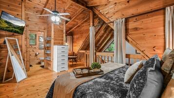 A cozy wooden cabin bedroom with a bed, a ceiling fan, a TV, and a small desk area.