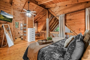 A cozy wooden cabin bedroom with a bed, a ceiling fan, a TV, and a small desk area.