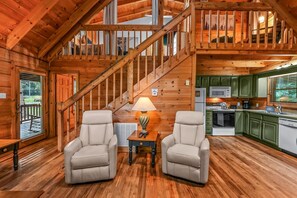 Ein Interieur einer Holzhütte mit Treppe, zwei Sesseln, einem Tisch mit Lampe und einer Küchenzeile.