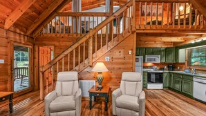 A wooden cabin interior with a staircase, two chairs, a table with a lamp, a kitchen area, and a view through a window. - Willow Creek 2 Bedroom Cabin by RedAwning (Townsend)