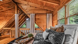 A cozy wooden cabin interior with a loft bedroom, a sofa with cushions, a tray with mugs and a plant, and a window with blinds. - Willow Creek 2 Bedroom Cabin by RedAwning (Townsend)