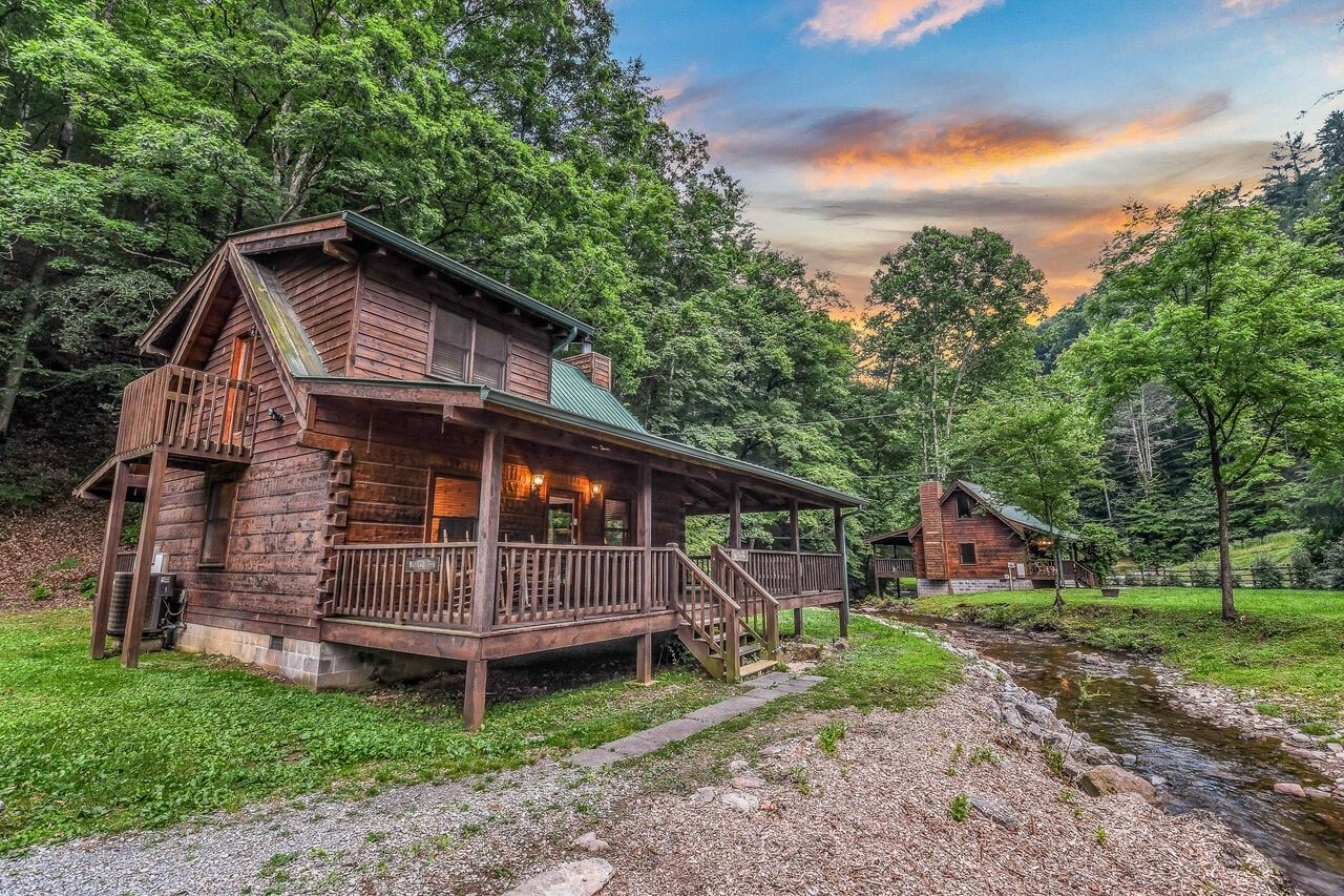 A rustic cabin with a porch and a small staircase, surrounded by lush greenery and a stream.