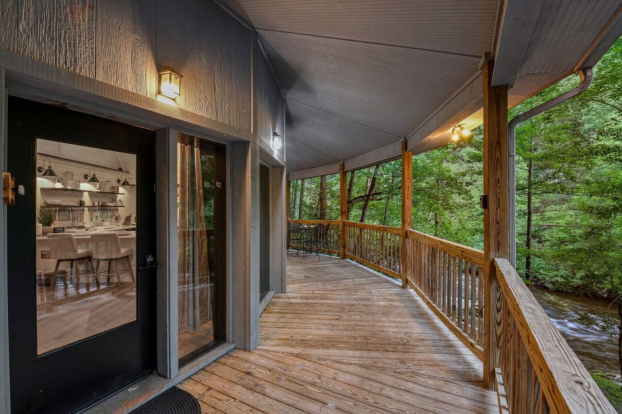 A wooden deck with a glass door leading to a kitchen area, surrounded by a forested landscape.