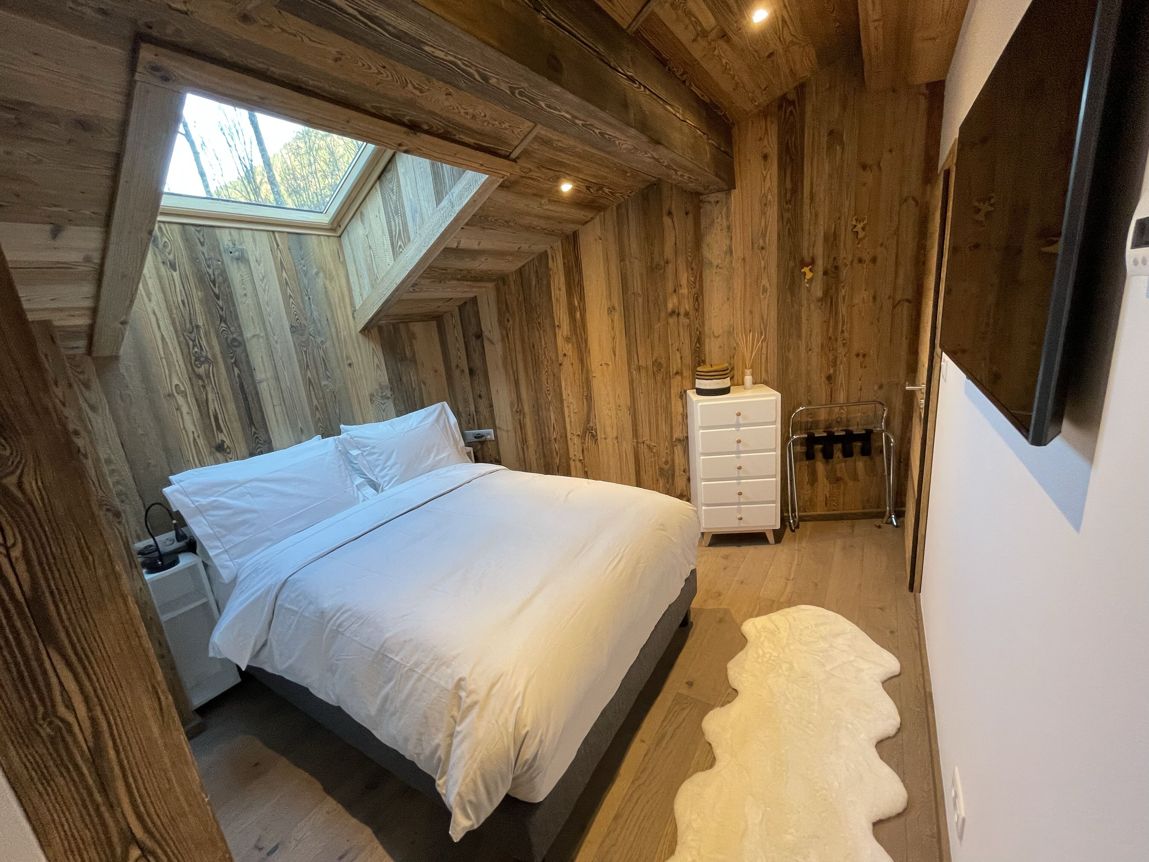 Une chambre avec un plafond en bois, une lucarne, un lit, une commode et un tapis blanc.