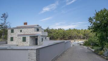 Un edificio de dos plantas con fachada de piedra y contraventanas verdes, junto a una zona pavimentada con muelle para barcos.